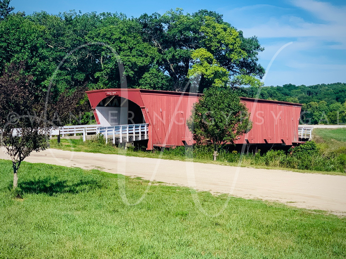 A photo of the Hogback Covered Bridge, a historic wooden bridge located in Madison County, Iowa. The bridge is surrounded by lush green trees and foliage, and features a peaked red roof with wooden beams supporting the structure. The bridge spans a calm river below.
