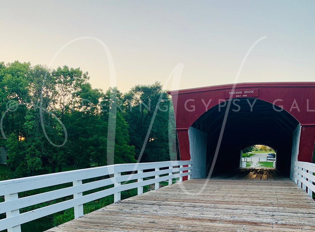 The Roseman Covered Bridge stands tall over a river, with a wooden roof and sides, surrounded by trees and greenery.